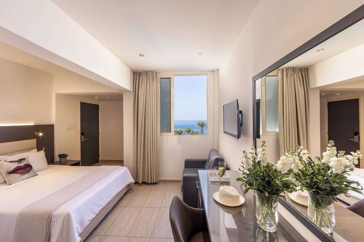 A hotel room with a bed, a sofa, a dining table with a vase of flowers, a TV, and a window with curtains.