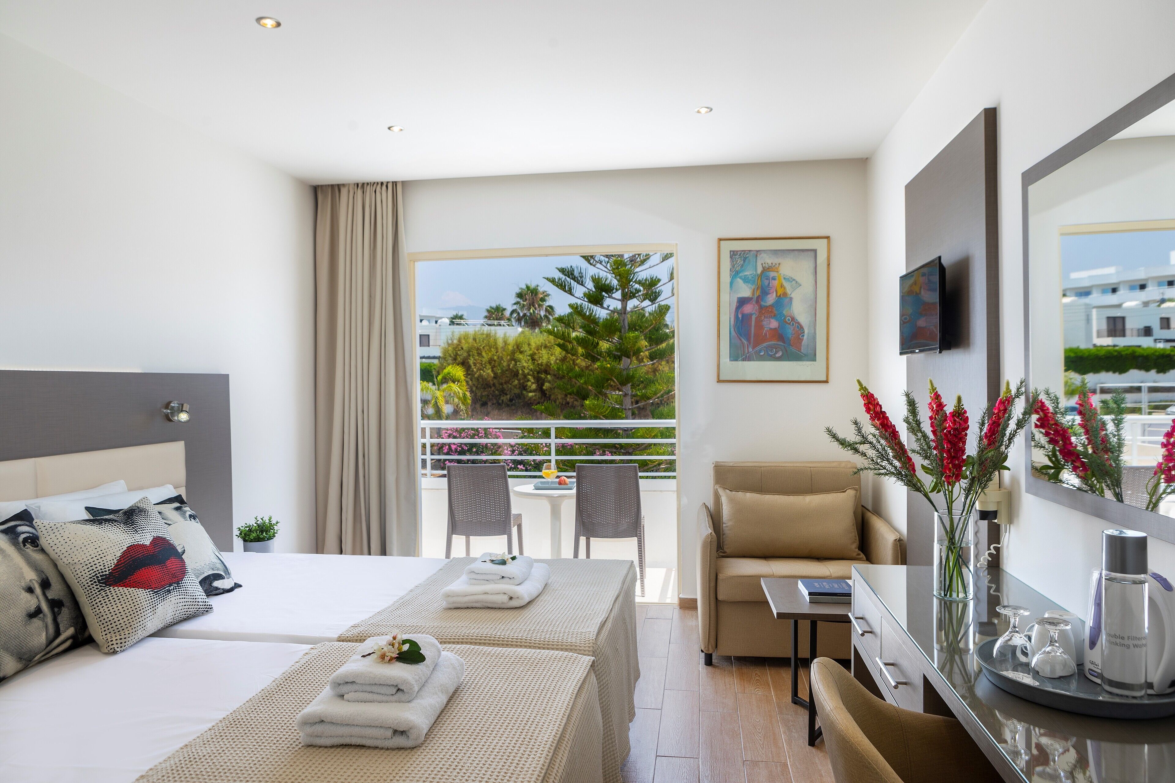 Habitación de hotel con cama, sofá, balcón con mesa y sillas, y vista a zonas verdes y edificios.