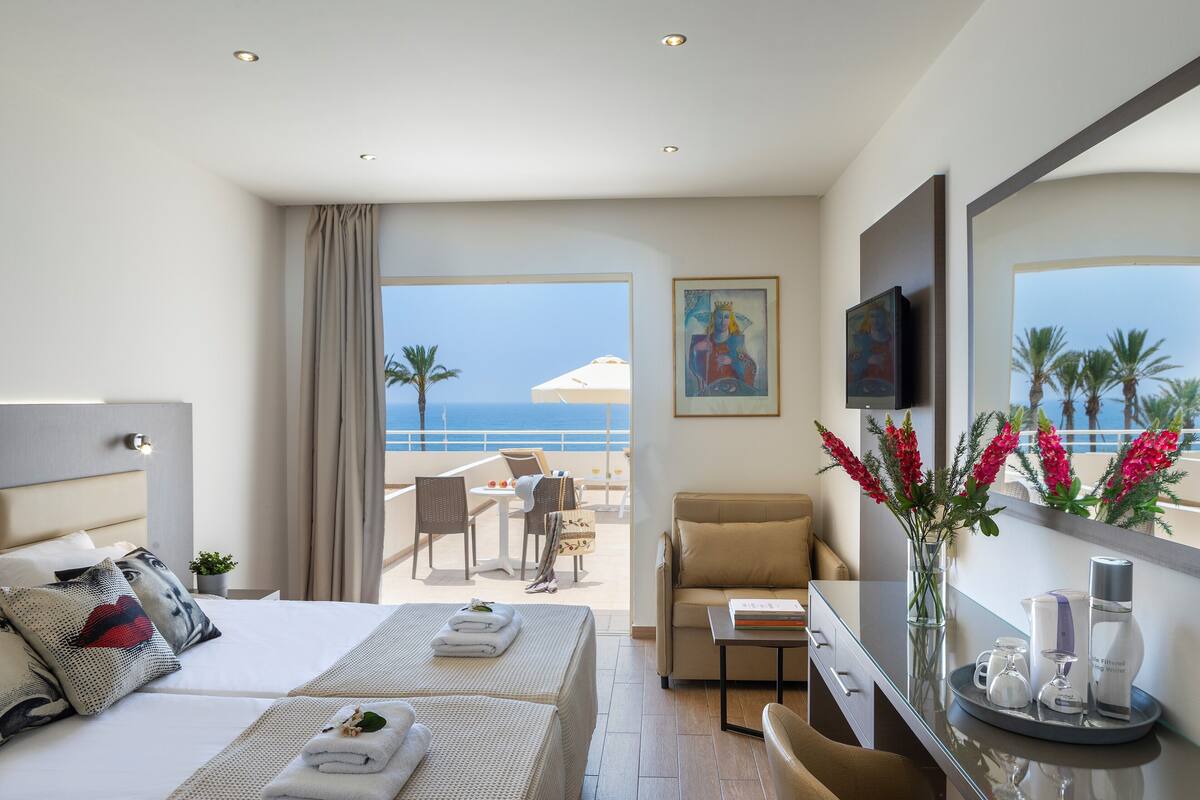 A hotel room with two beds, a balcony with a view of the sea, a dining area, and a bathroom with a large mirror.