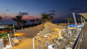 An outdoor dining area with tables and chairs, palm trees, and a view of the sea at dusk.