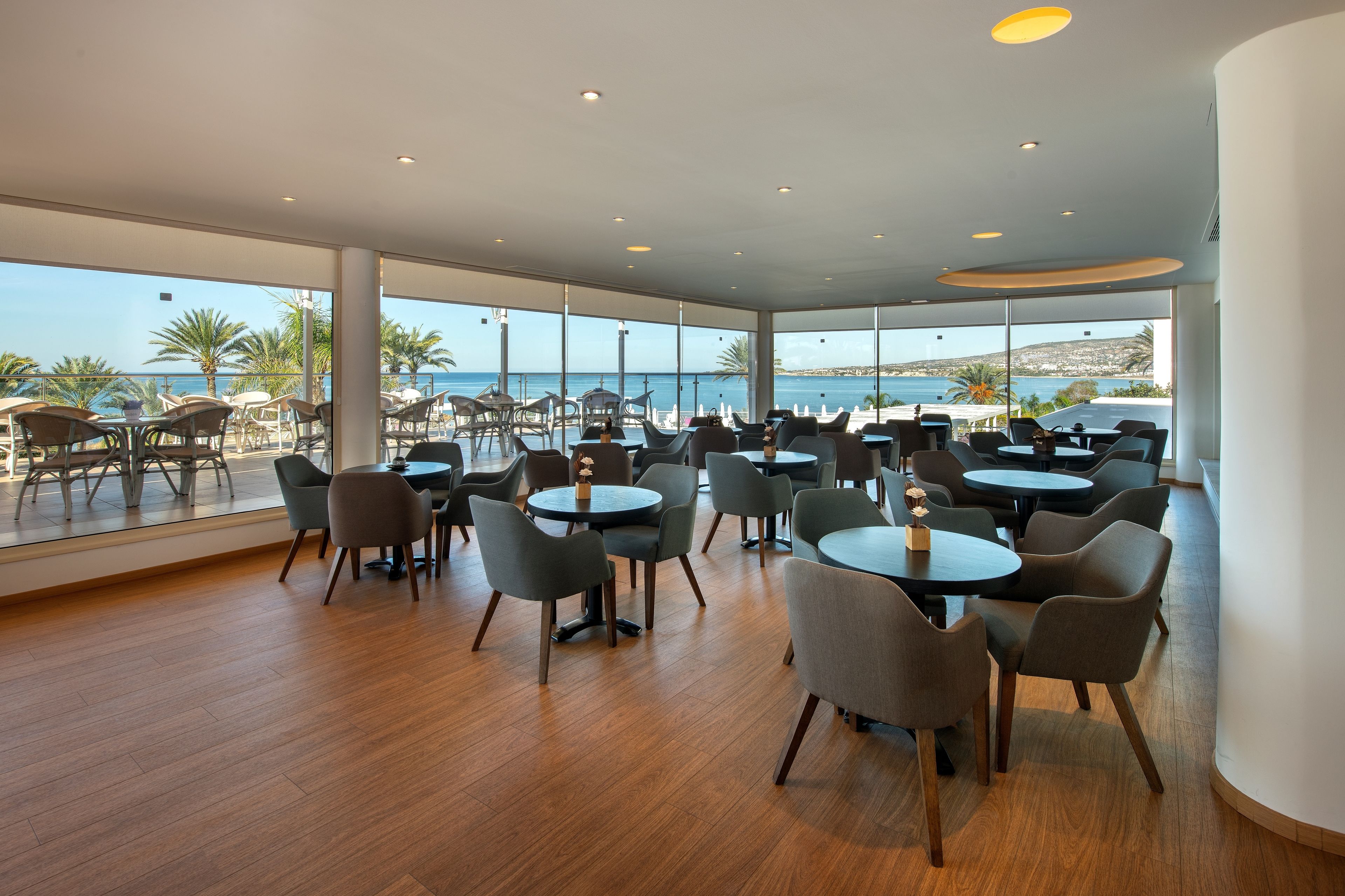Un amplio comedor interior con varias mesas y sillas, grandes ventanales que ofrecen vista al mar y palmeras.