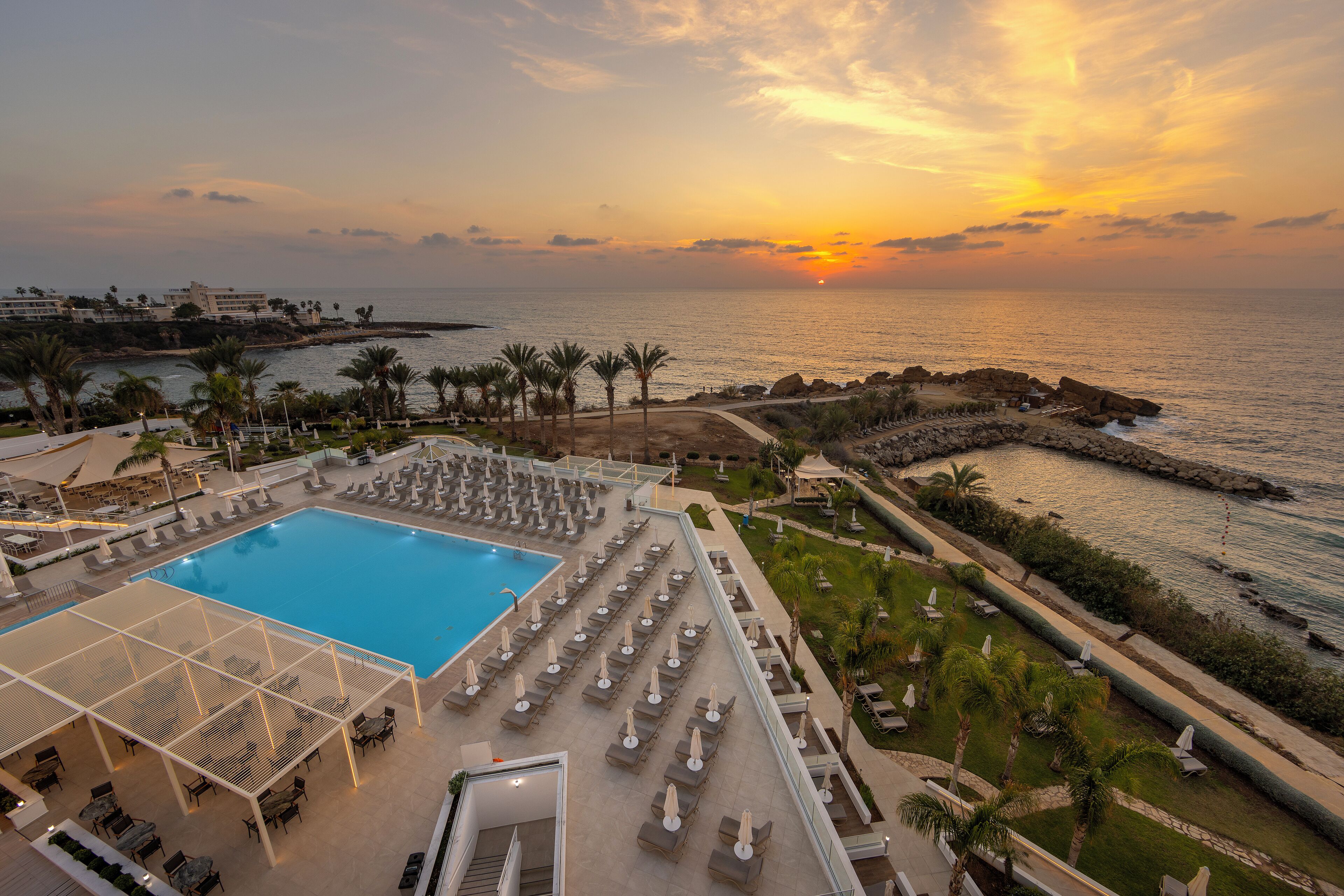 A resort with a large pool, sun loungers, and a view of the sunset over the ocean.