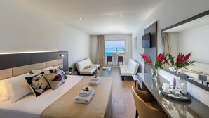 A modern hotel room with a bed, two sofas, a dining area, and a view of the sea. - Queen's Bay Hotel (Kissonerga)