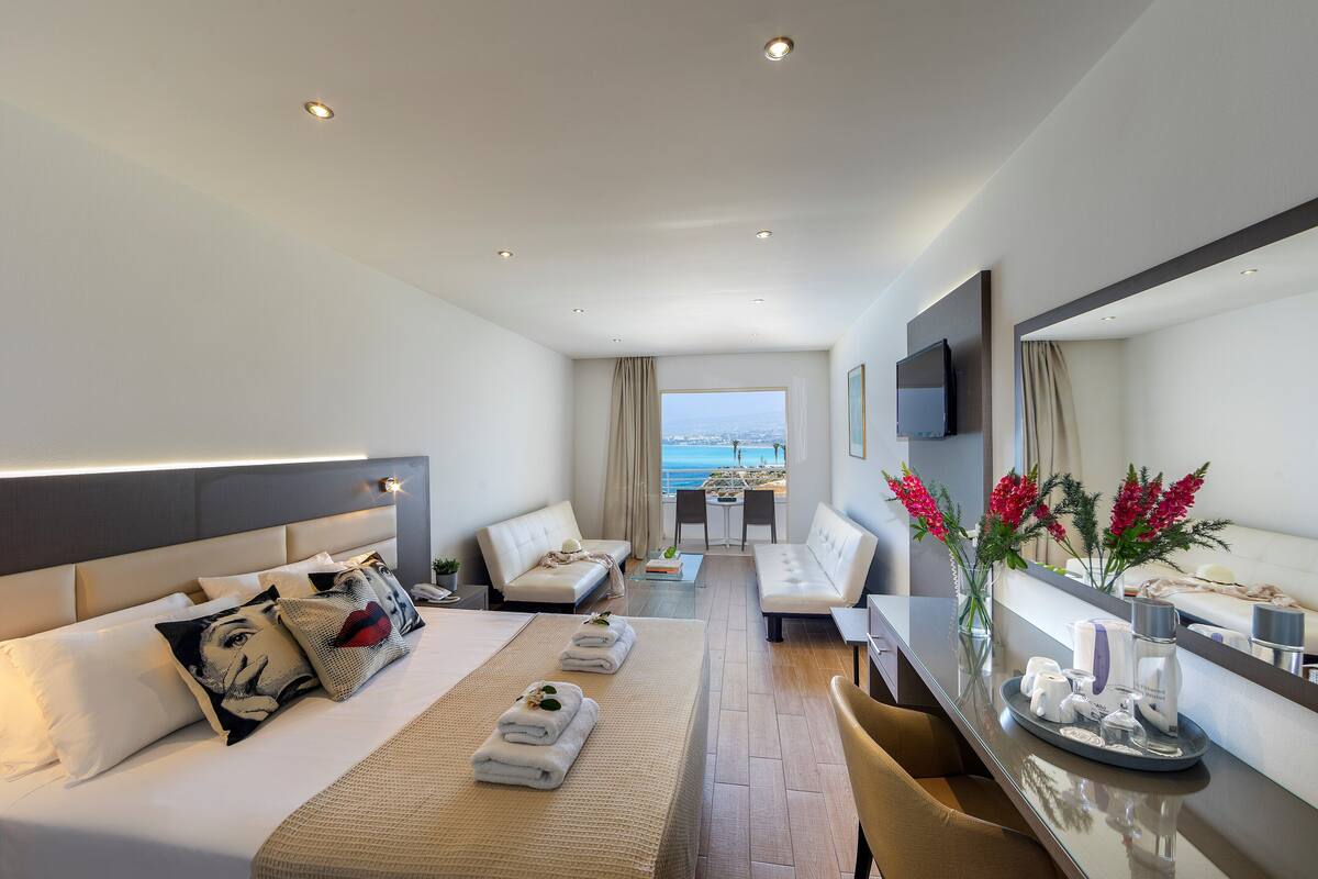 A modern hotel room with a large bed, a sofa, a dining area, and a view of the sea.
