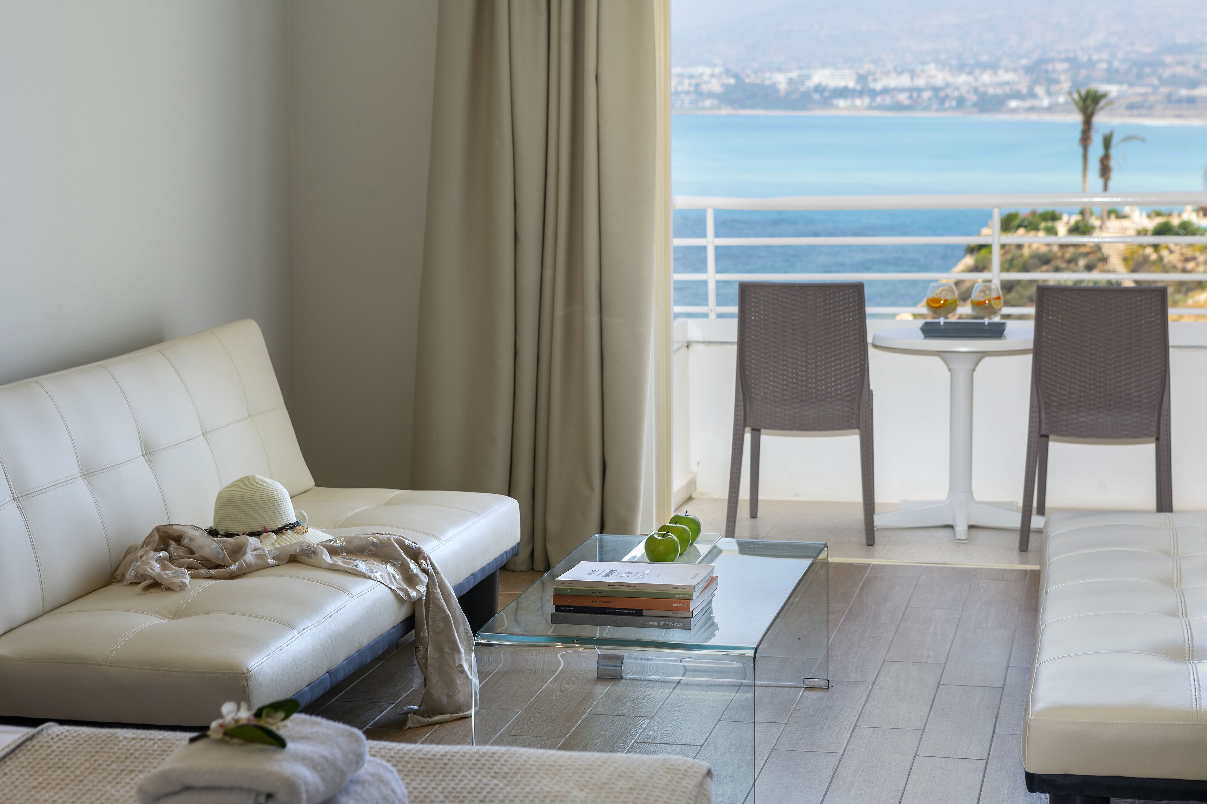 A modern living room with a white sofa, a glass coffee table with books and a green apple, and a view of the sea.