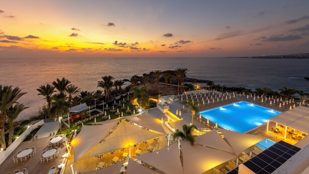 A resort with a swimming pool, outdoor seating, and palm trees. - Queen's Bay Hotel (Kissonerga)