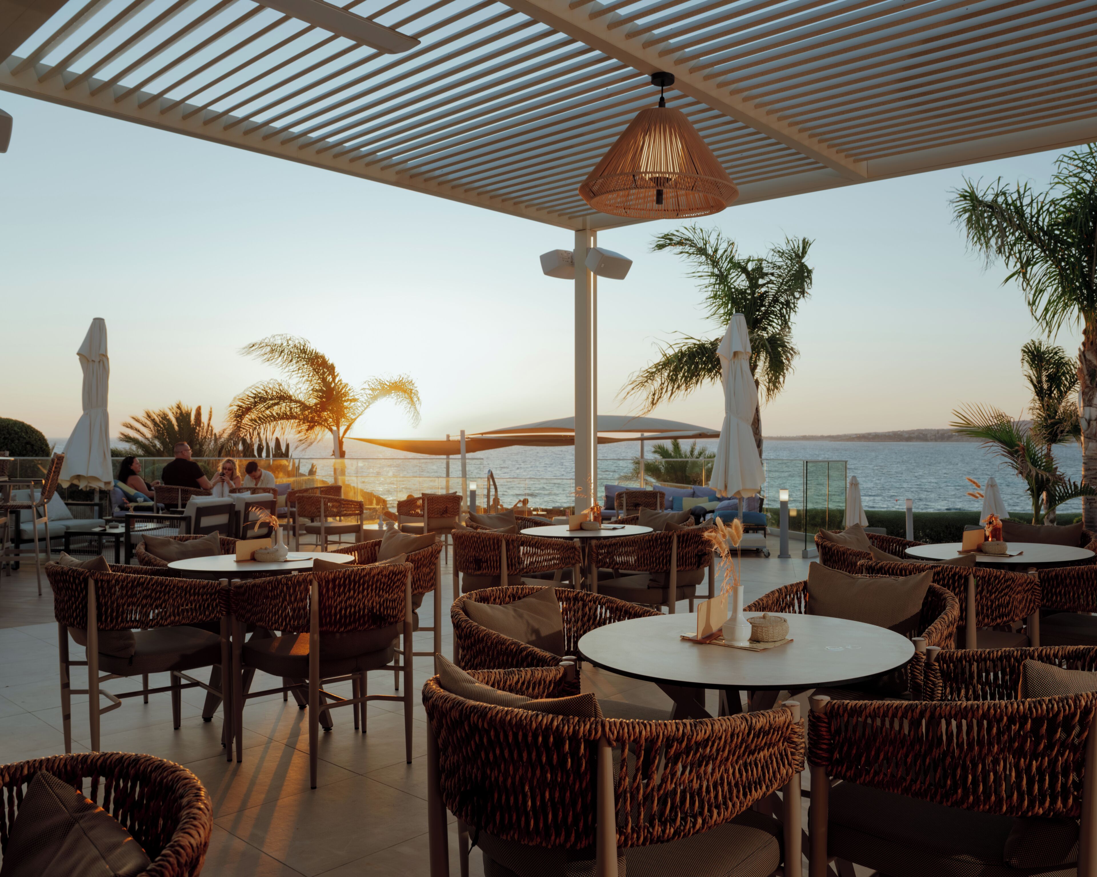 An outdoor seating area with wicker furniture and umbrellas, overlooking a body of water.