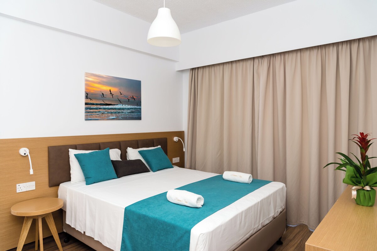 A neatly made bed with a teal bedspread and pillows, a white wall with a sunset painting, a wooden nightstand with a lamp, and a potted plant on a wooden table.