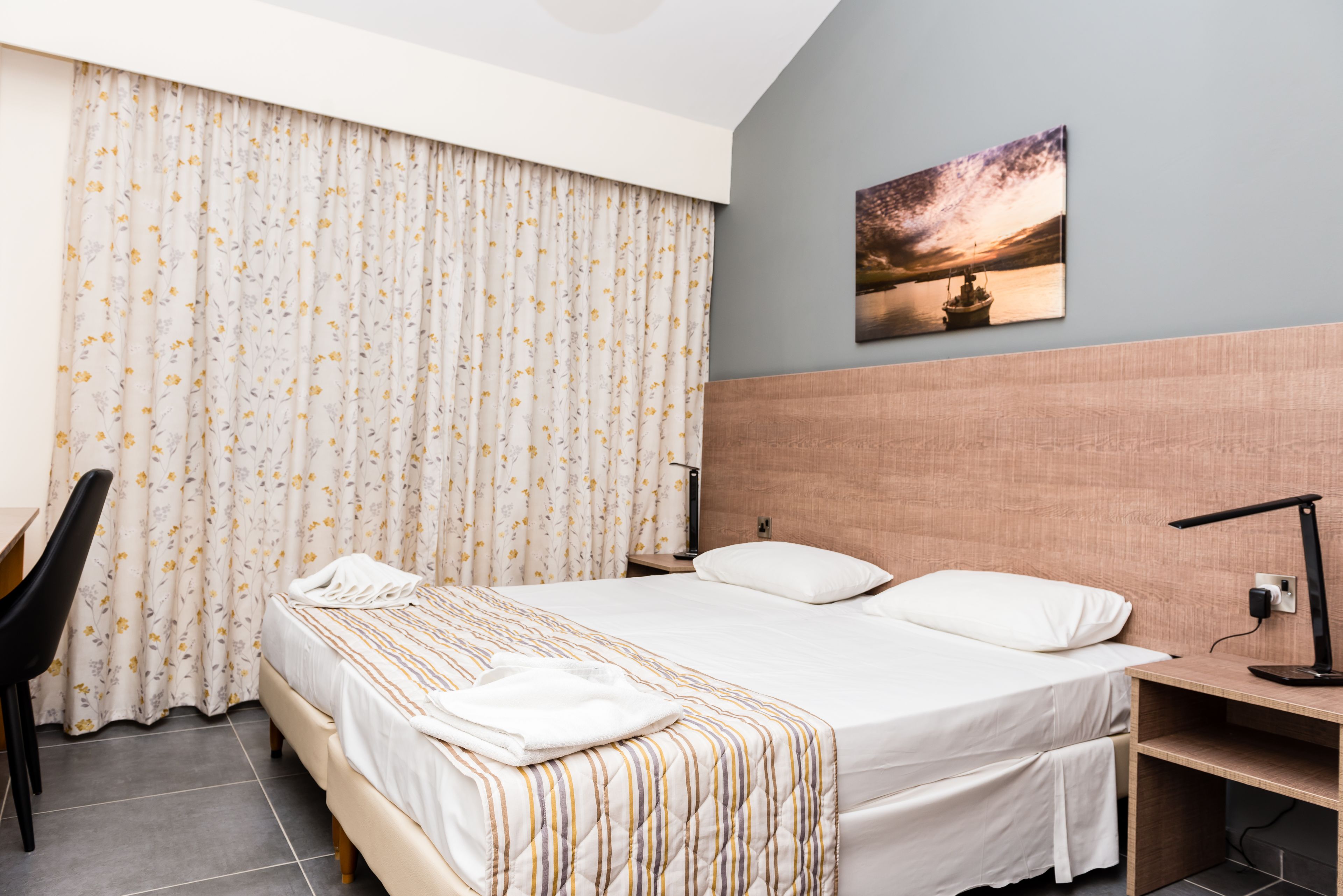 A hotel room with a bed, a desk, a chair, a nightstand, and a painting on the wall.