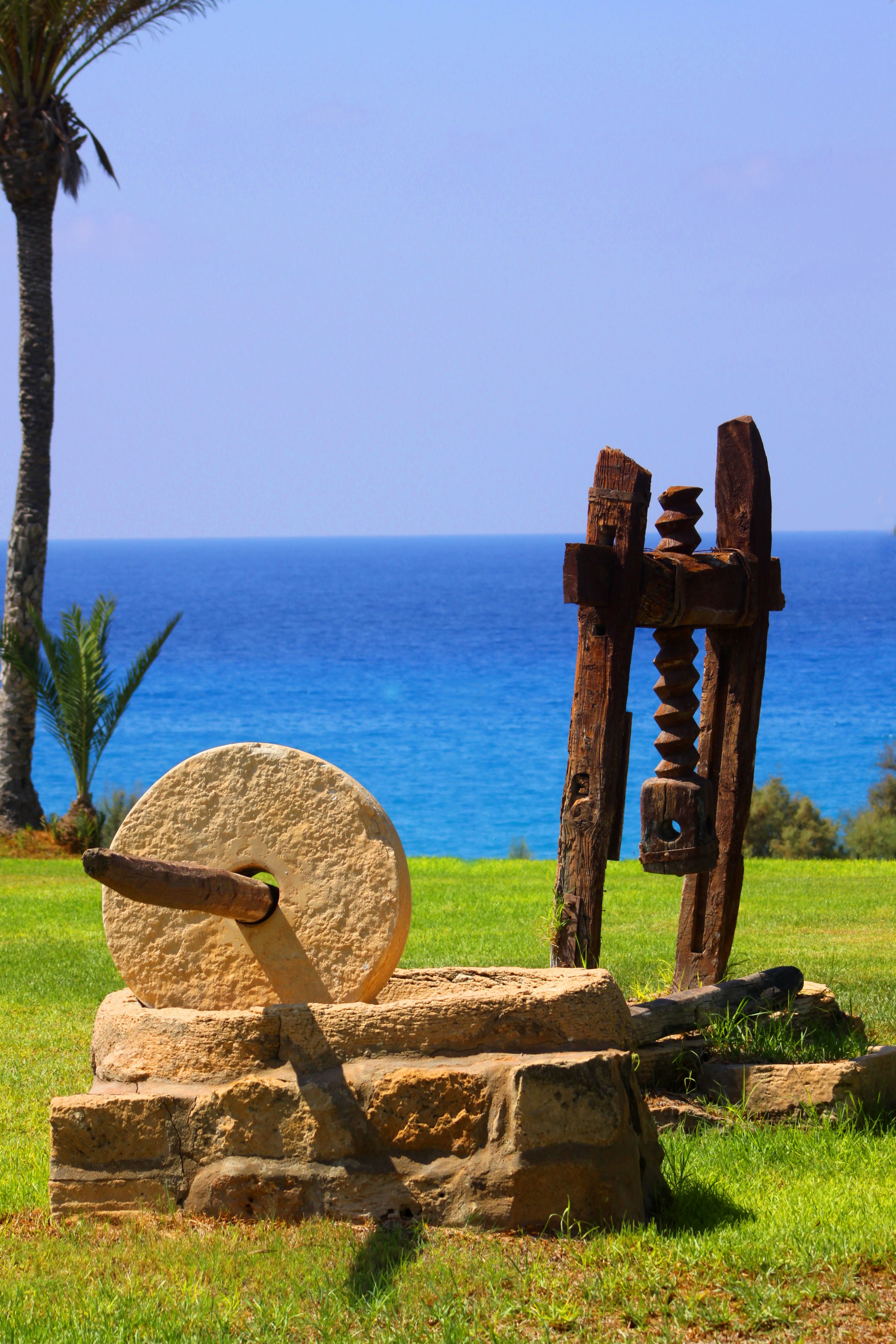A stone mill wheel and wooden mill components on a grassy hill with a view of the ocean.