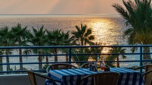 A seaside restaurant with tables and chairs, palm trees, and a view of the ocean at sunset. - Helios Bay Hotel Apartments (Chlorakas)