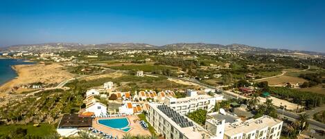 Vista aérea de un complejo turístico con piscina, varias edificaciones y una playa al fondo.