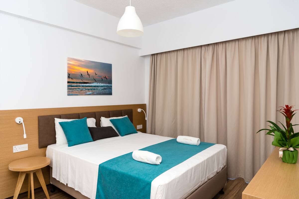 A hotel room with a bed, a nightstand, a lamp, a painting, and a plant.