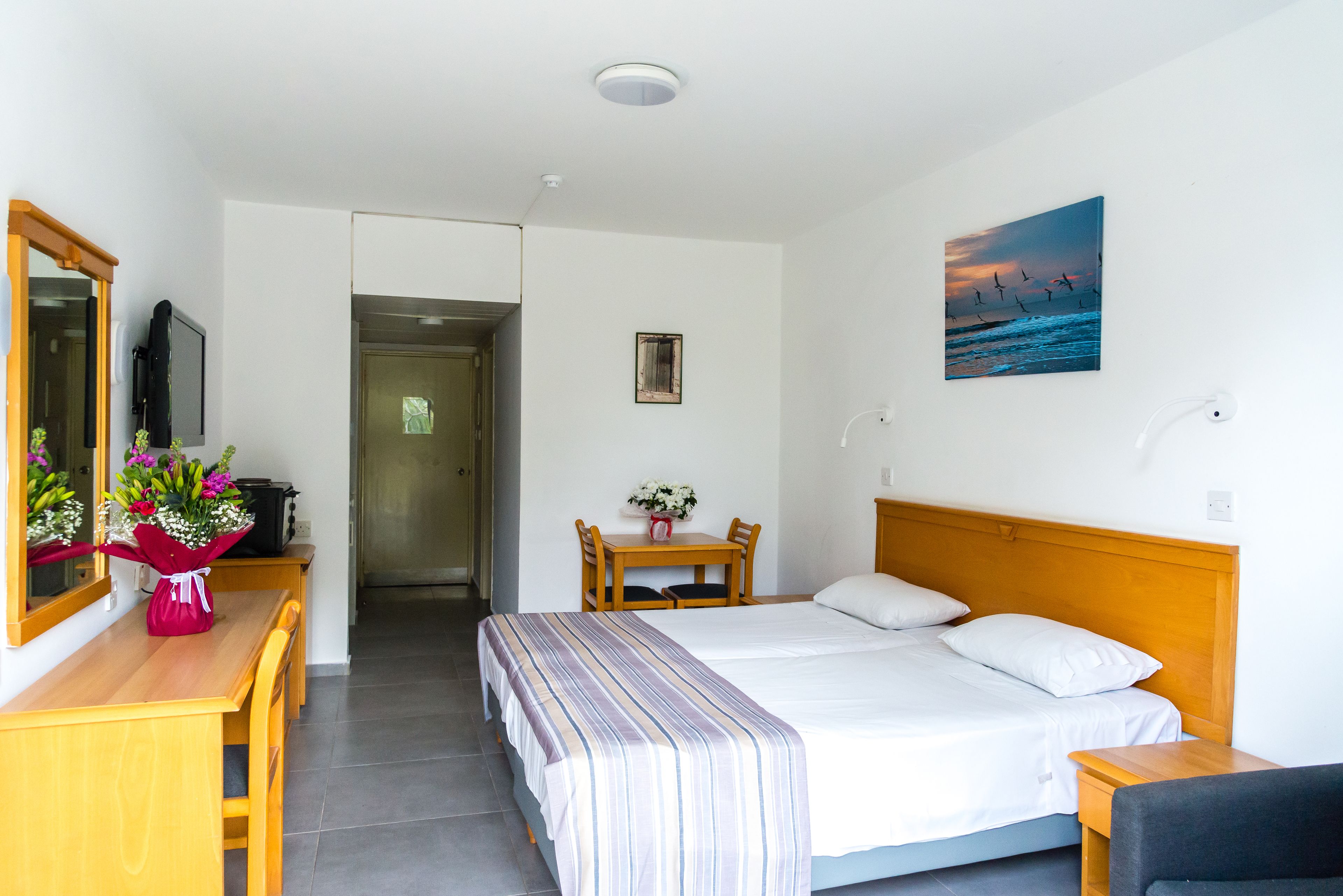 Ett hotellrum med en säng, ett skrivbord med en stol, ett litet bord med en vas med blommor och en tavla på väggen.