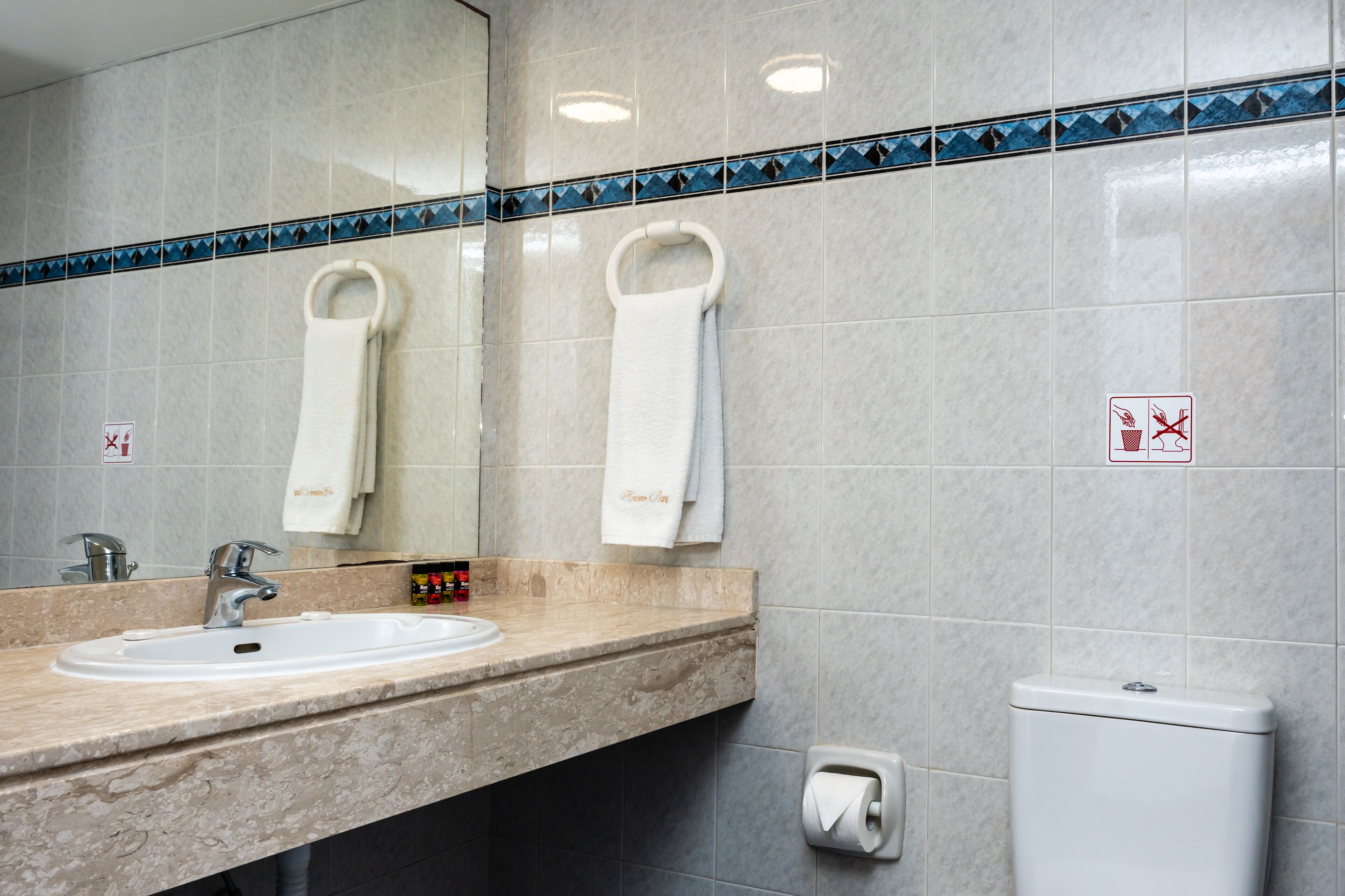 A bathroom with a sink, mirror, towel rings, and a toilet.