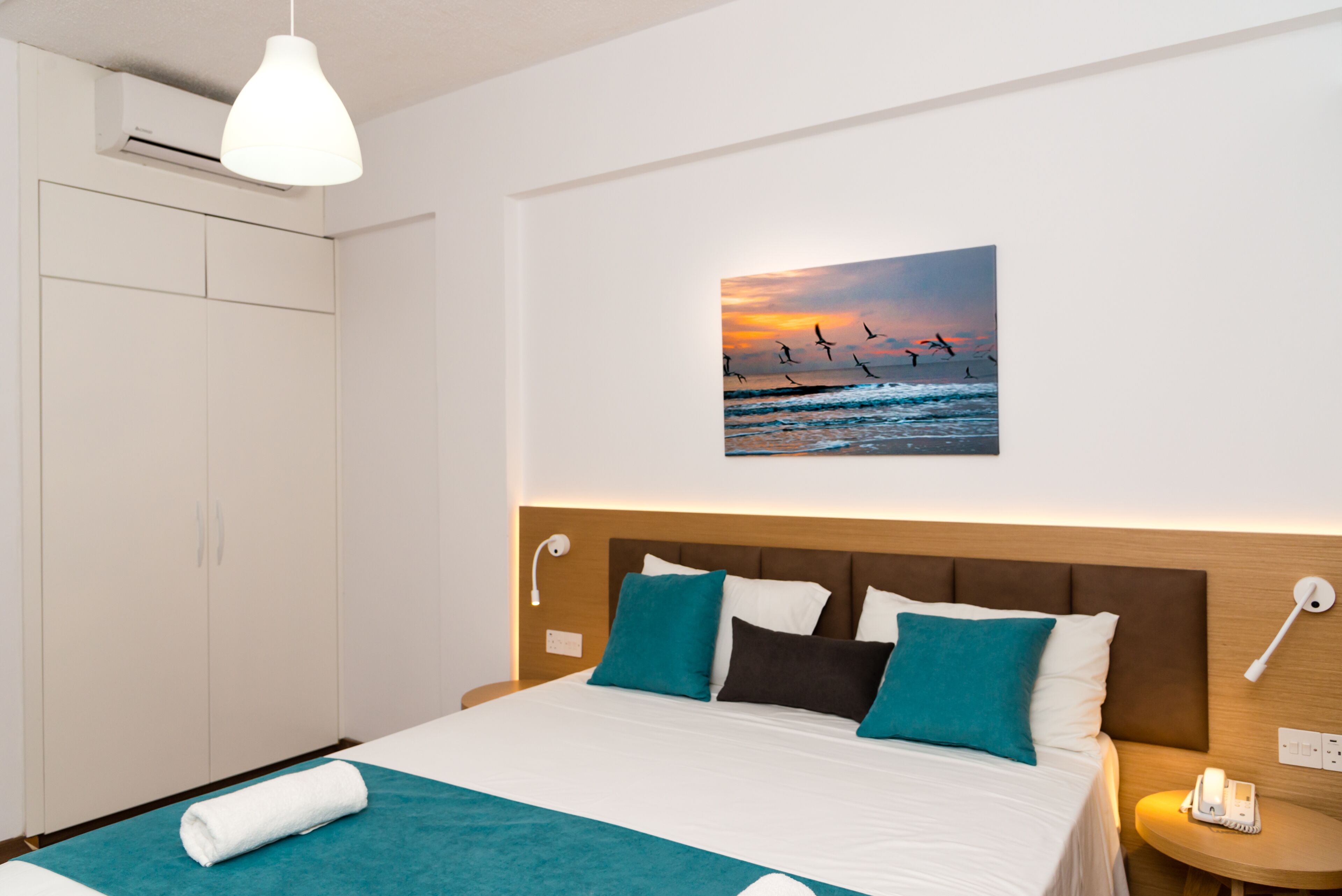 A hotel room with a bed, a painting of birds at sunset, a white lamp, and a wooden nightstand.