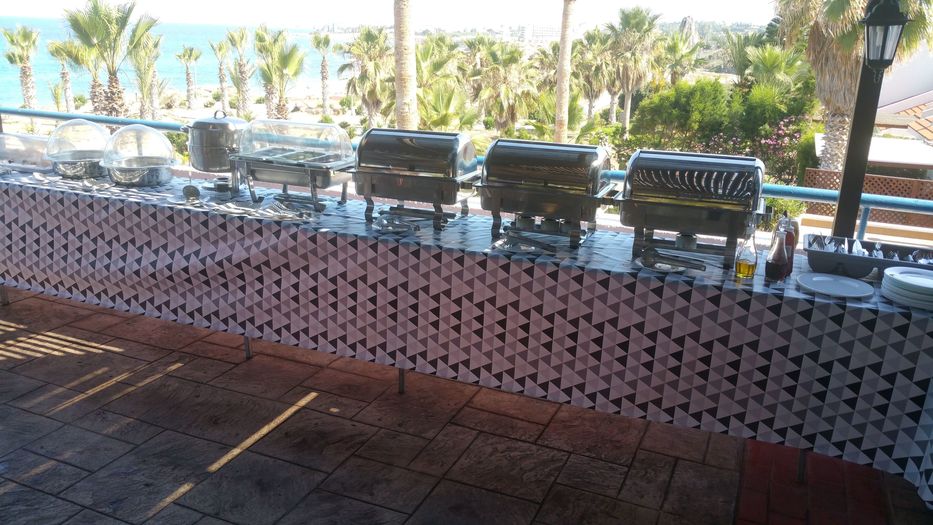 Montaje de bufé con varias parrillas y platos de servicio, un mantel estampado y vista al océano y palmeras al fondo.