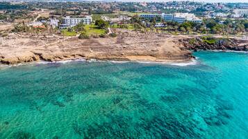 Luchtbeeld van een kustgebied met helder turquoise water, een rotsachtige kustlijn en een combinatie van woongebouwen en begroeiing.