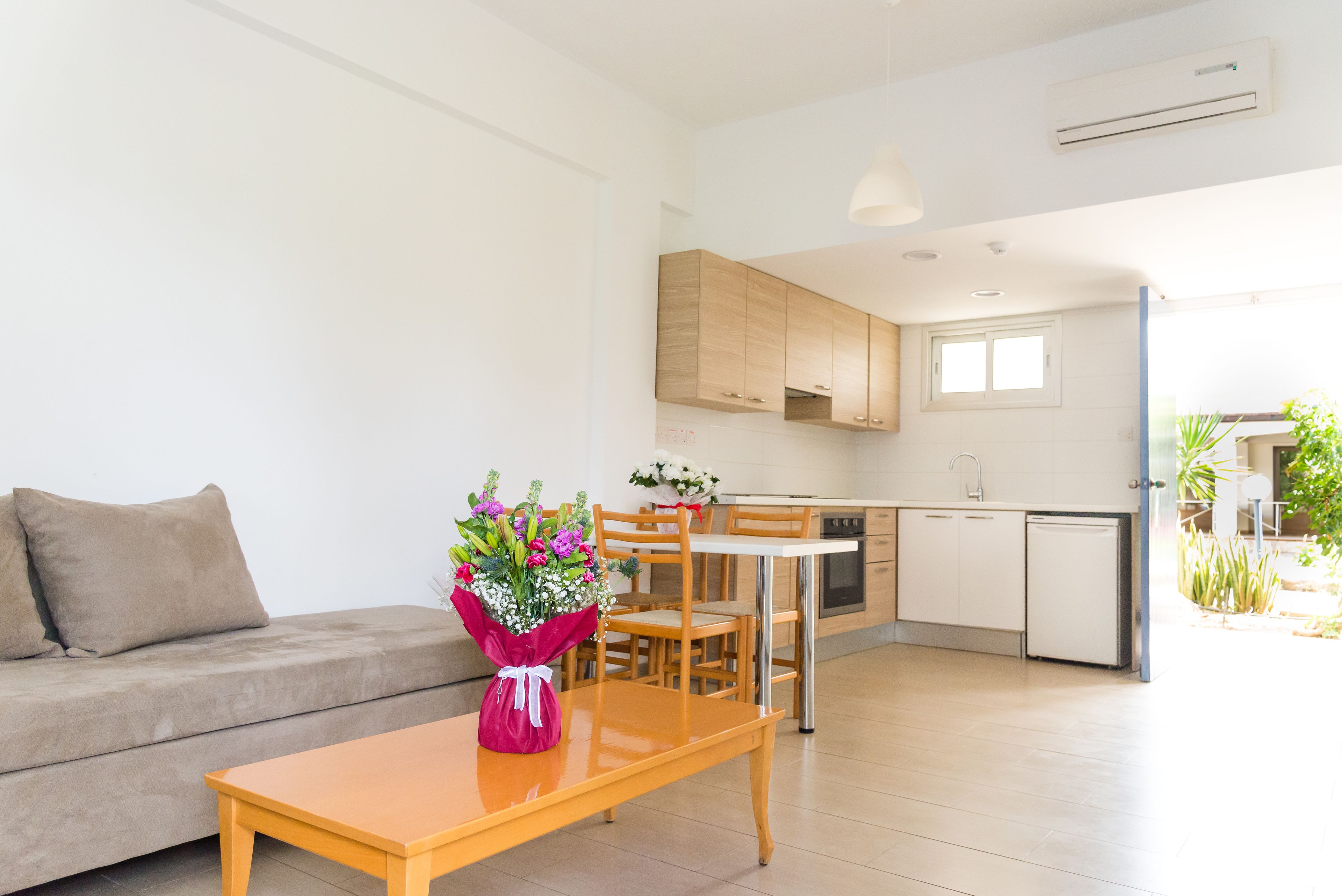 A modern living room with a sofa, a wooden table, and a bouquet of flowers. There is a kitchen area with cabinets and a refrigerator.