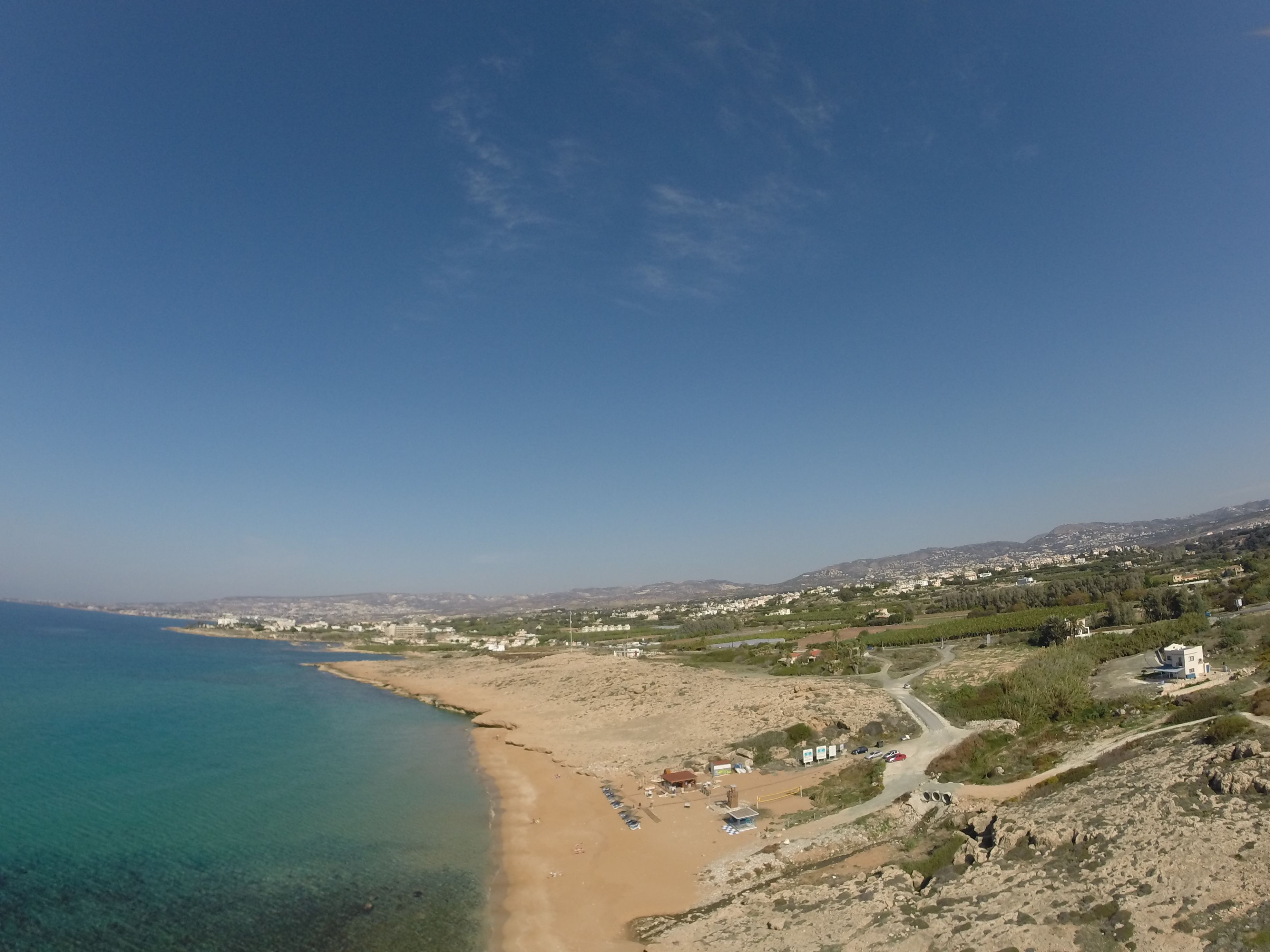 A coastal landscape with a clear view of the beach, a road leading to a building, and distant hills.