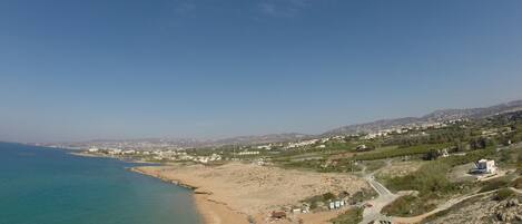 A coastal landscape with a clear view of the beach, a road leading to a building, and distant hills.