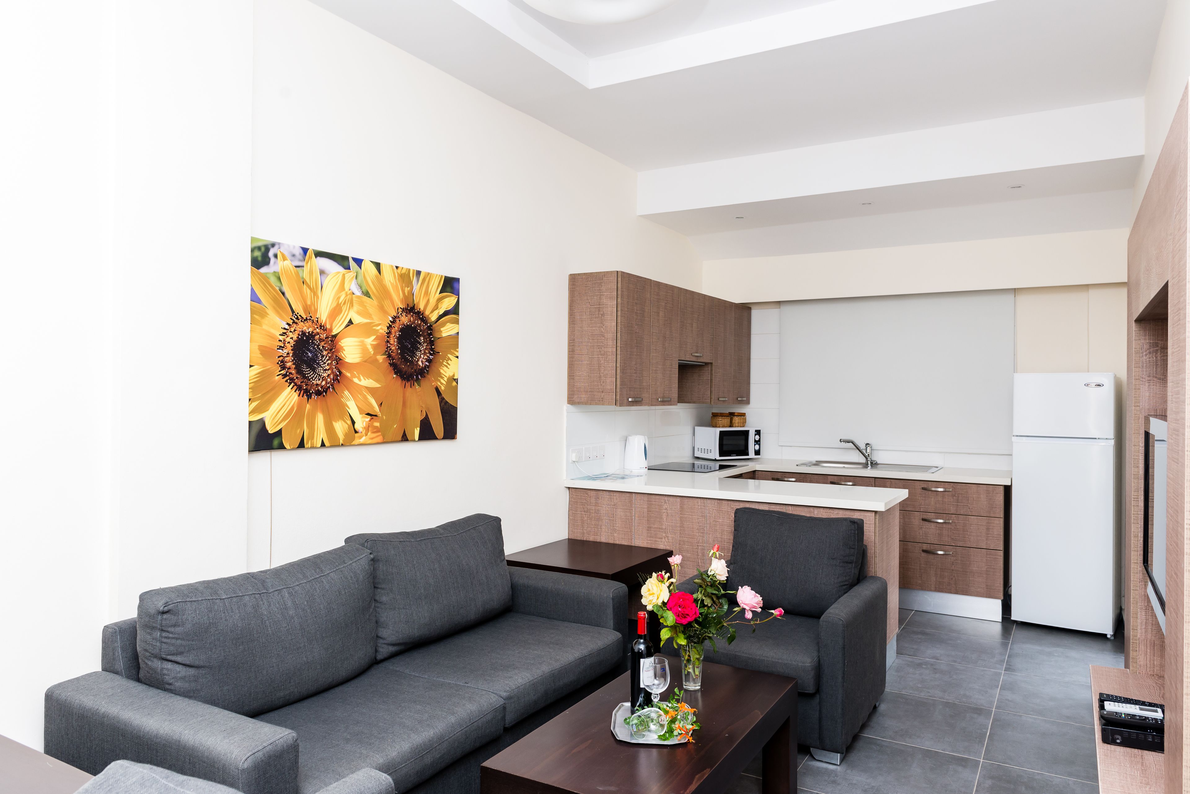 Un appartement moderne avec un canapé gris, une table basse ornée d’un vase de fleurs et un coin cuisine équipé d’un four à micro-ondes, d’un évier et de placards.