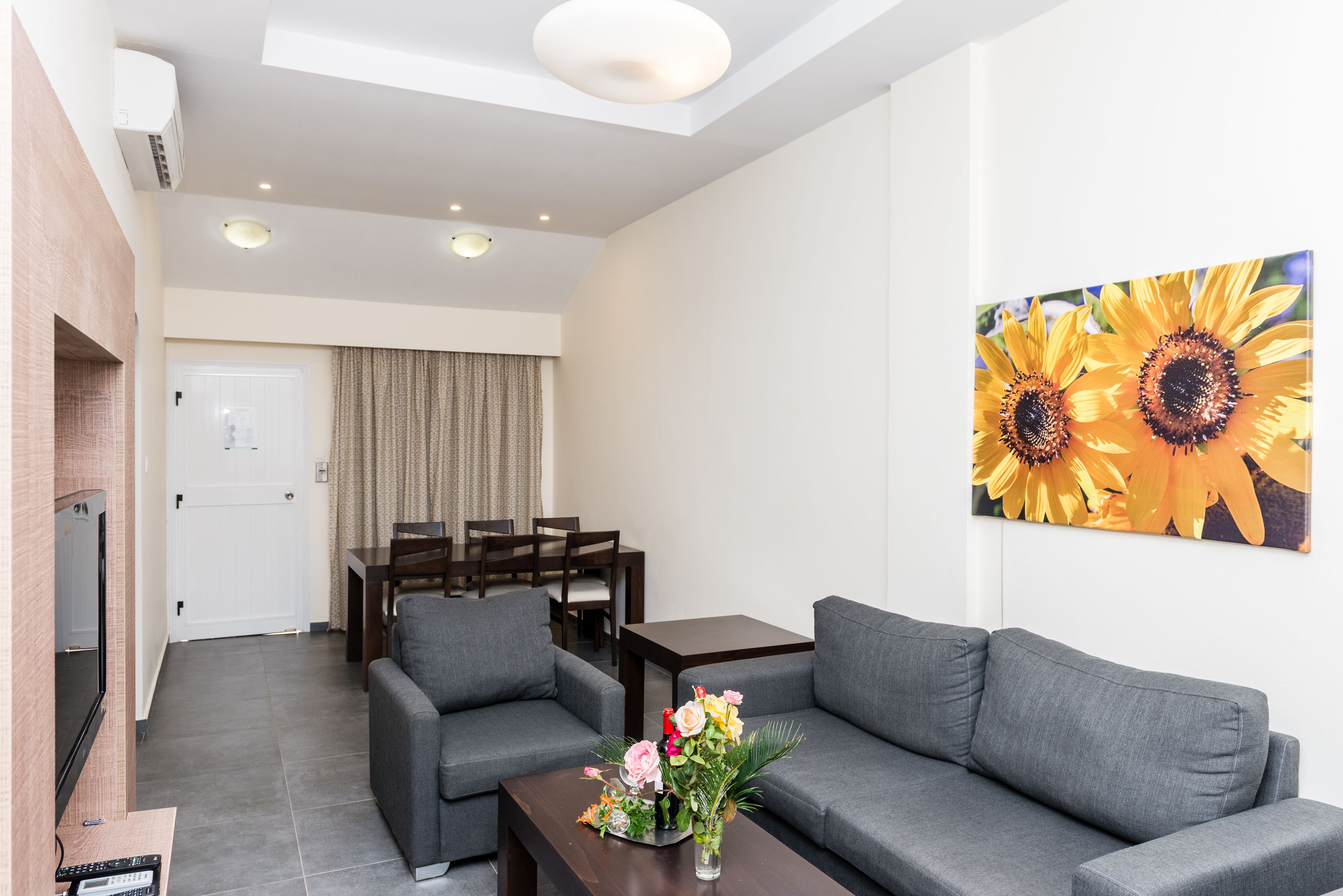 Una sala de estar moderna con un sofá gris, una mesa de centro con flores, un televisor y un comedor con mesa y sillas.