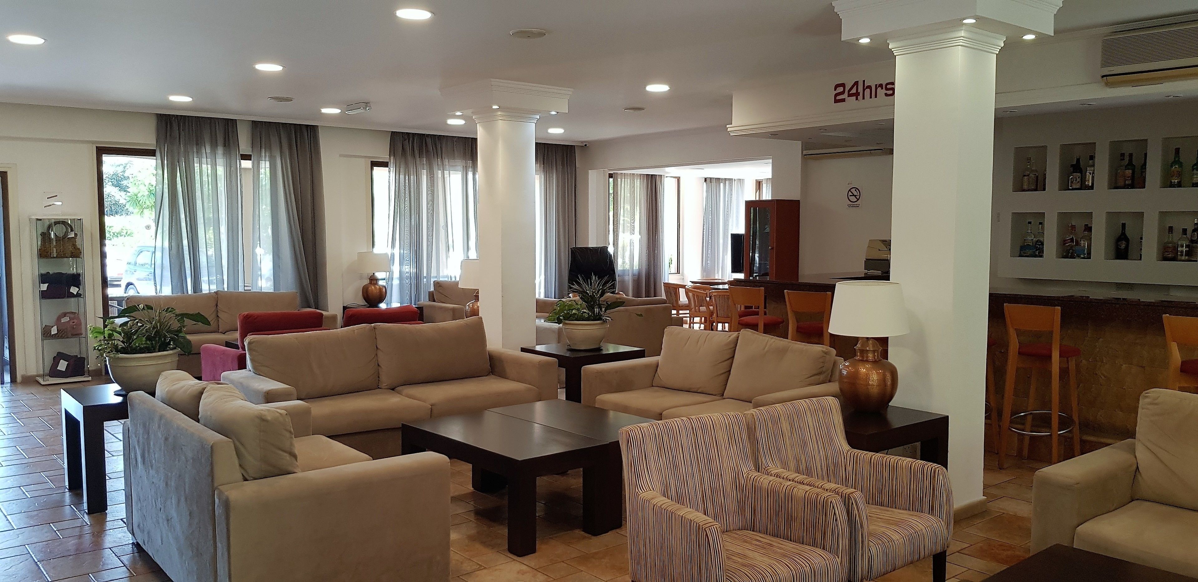 A spacious hotel lobby with beige sofas, a striped armchair, a coffee table, and a dining area with wooden chairs.