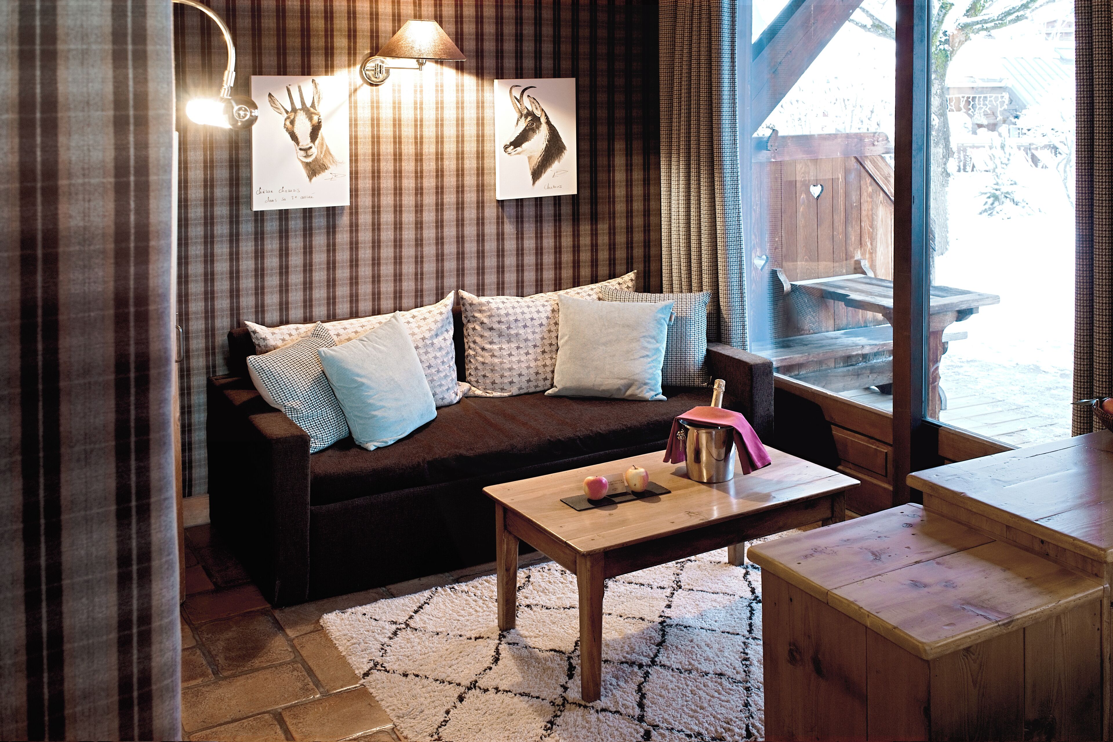 A cozy room with a brown sofa, patterned cushions, a wooden coffee table, and a view of a wooden deck outside.