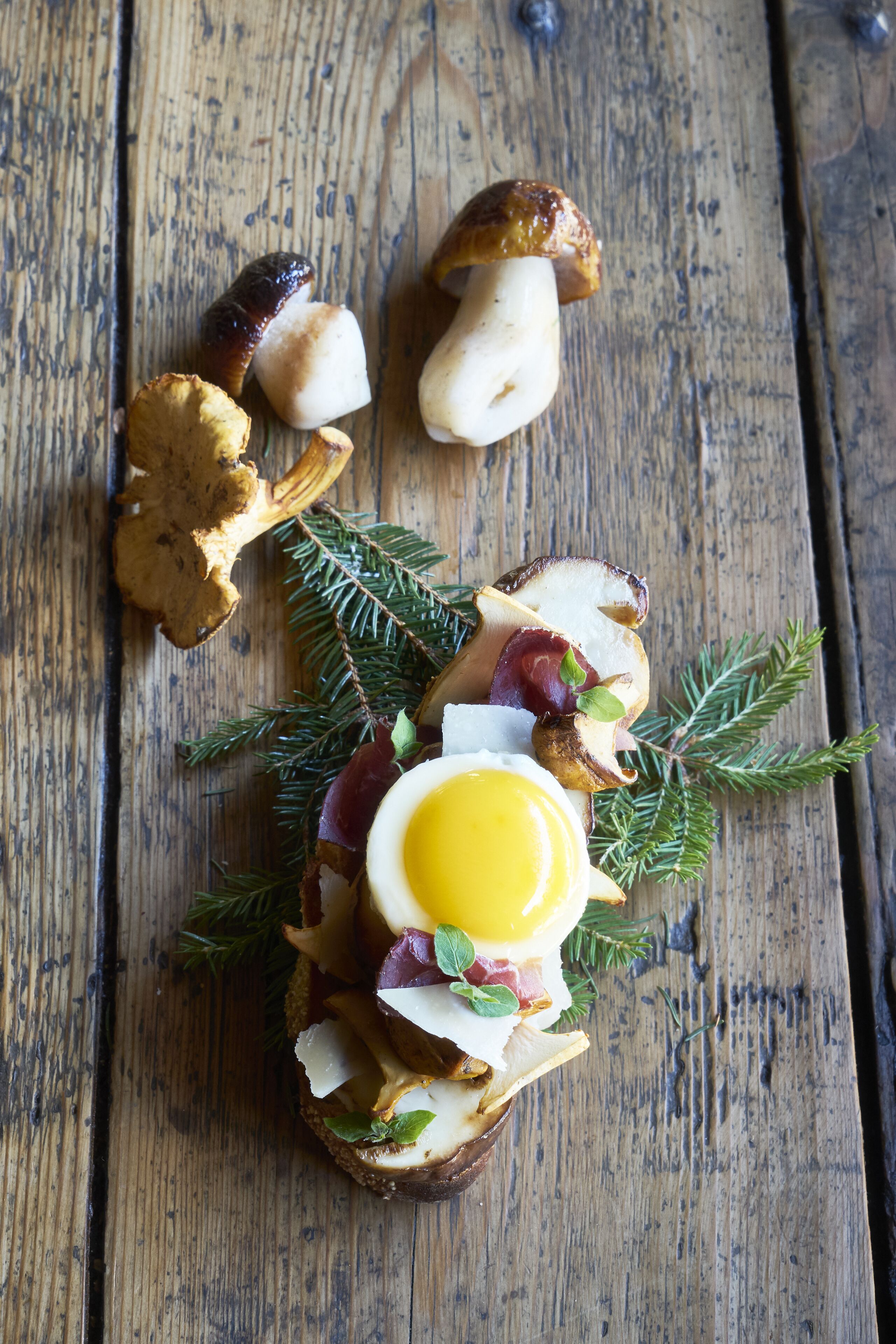 A rustic wooden table with a variety of food items, including a toasted bread with egg, mushrooms, and herbs.