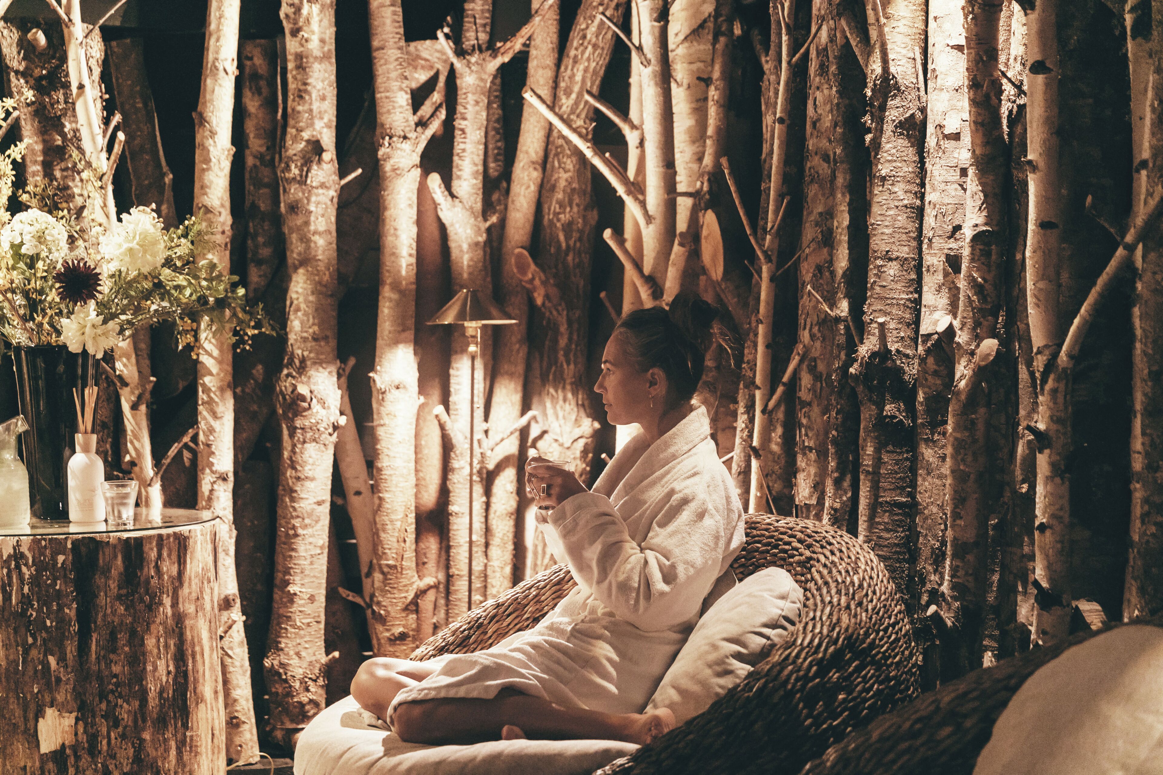 A person sitting in a wicker chair in a rustic setting with wooden elements and a table with a vase of flowers.