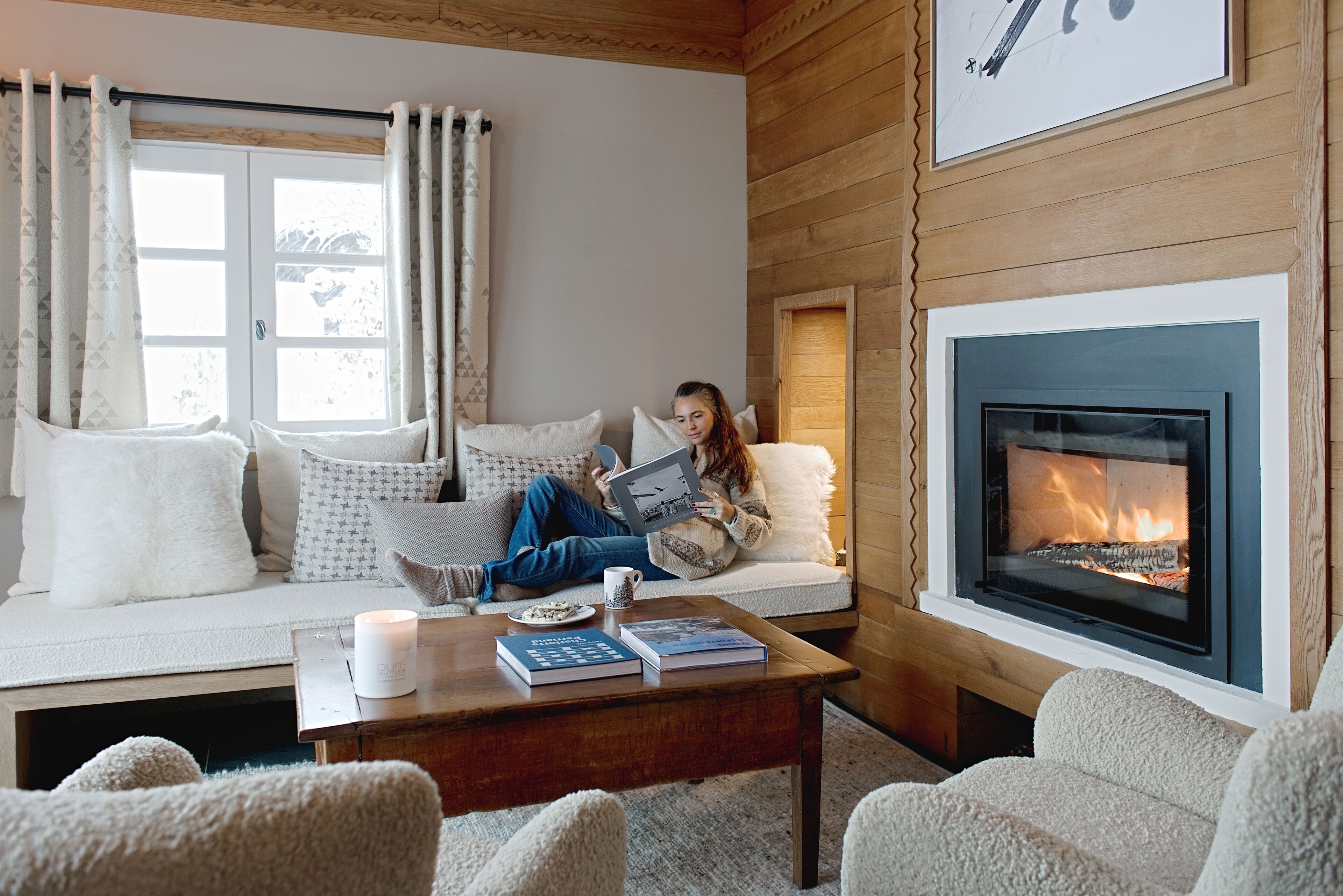 A cozy living room with a fireplace, a sofa with patterned cushions, and a coffee table with books and a candle.
