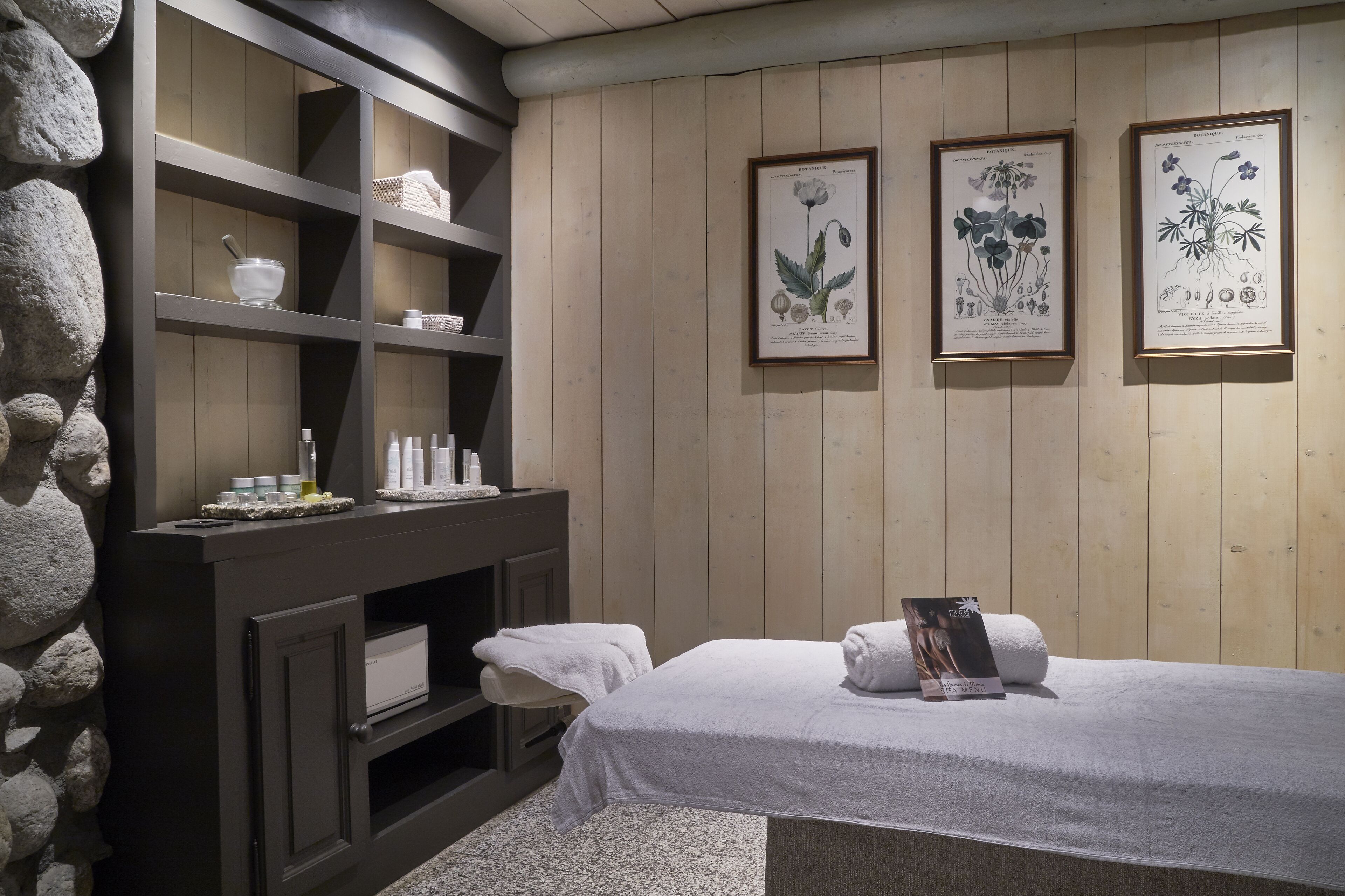 A spa room with a stone wall, wooden shelves, a massage table, and botanical illustrations on the wall.