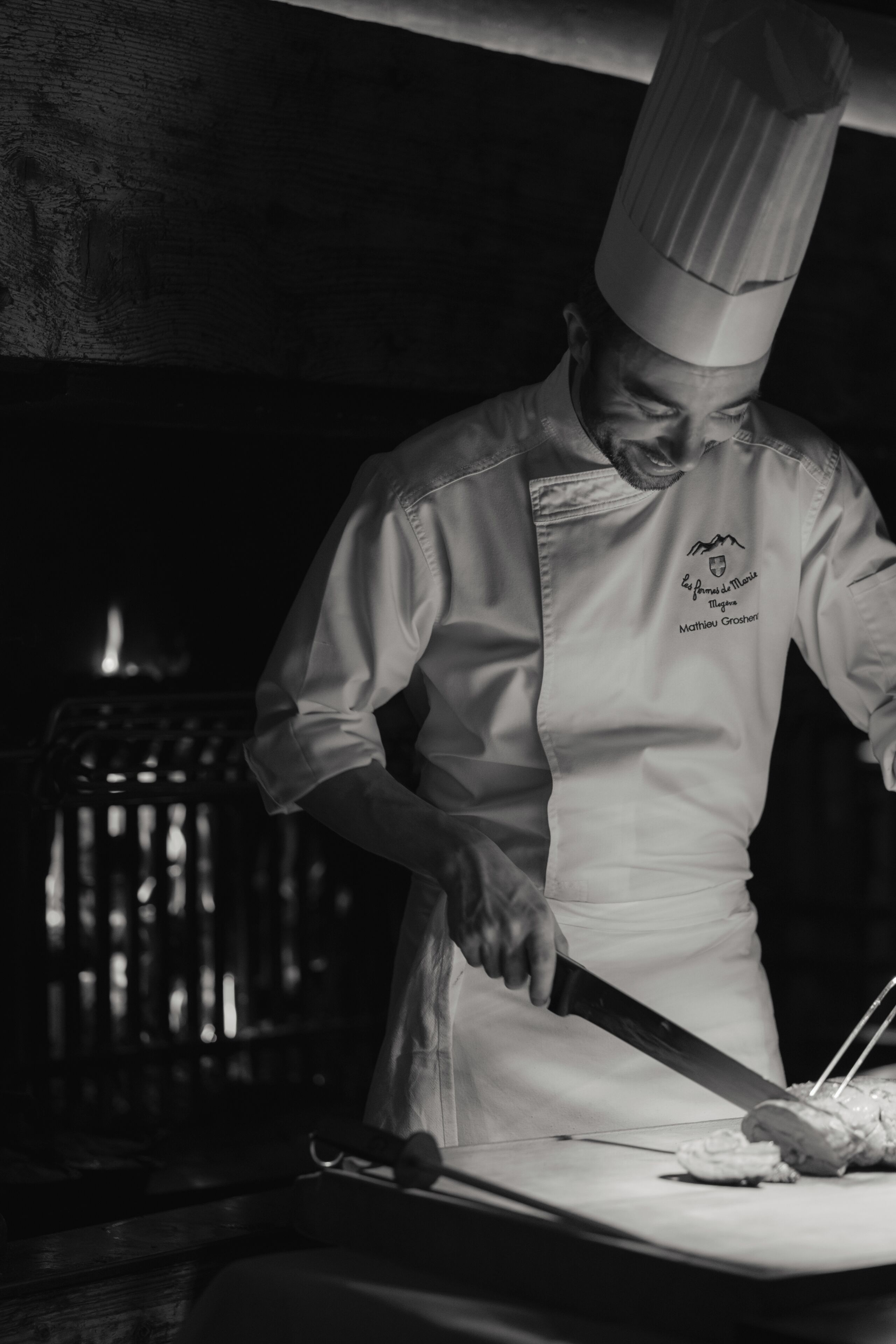 Un chef con uniforme blanco y un gorro alto, cortando comida sobre una tabla de cortar.