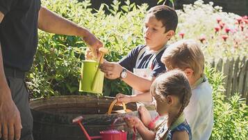 A man and children watering plants in a garden with a wooden barrel and a fence in the background.