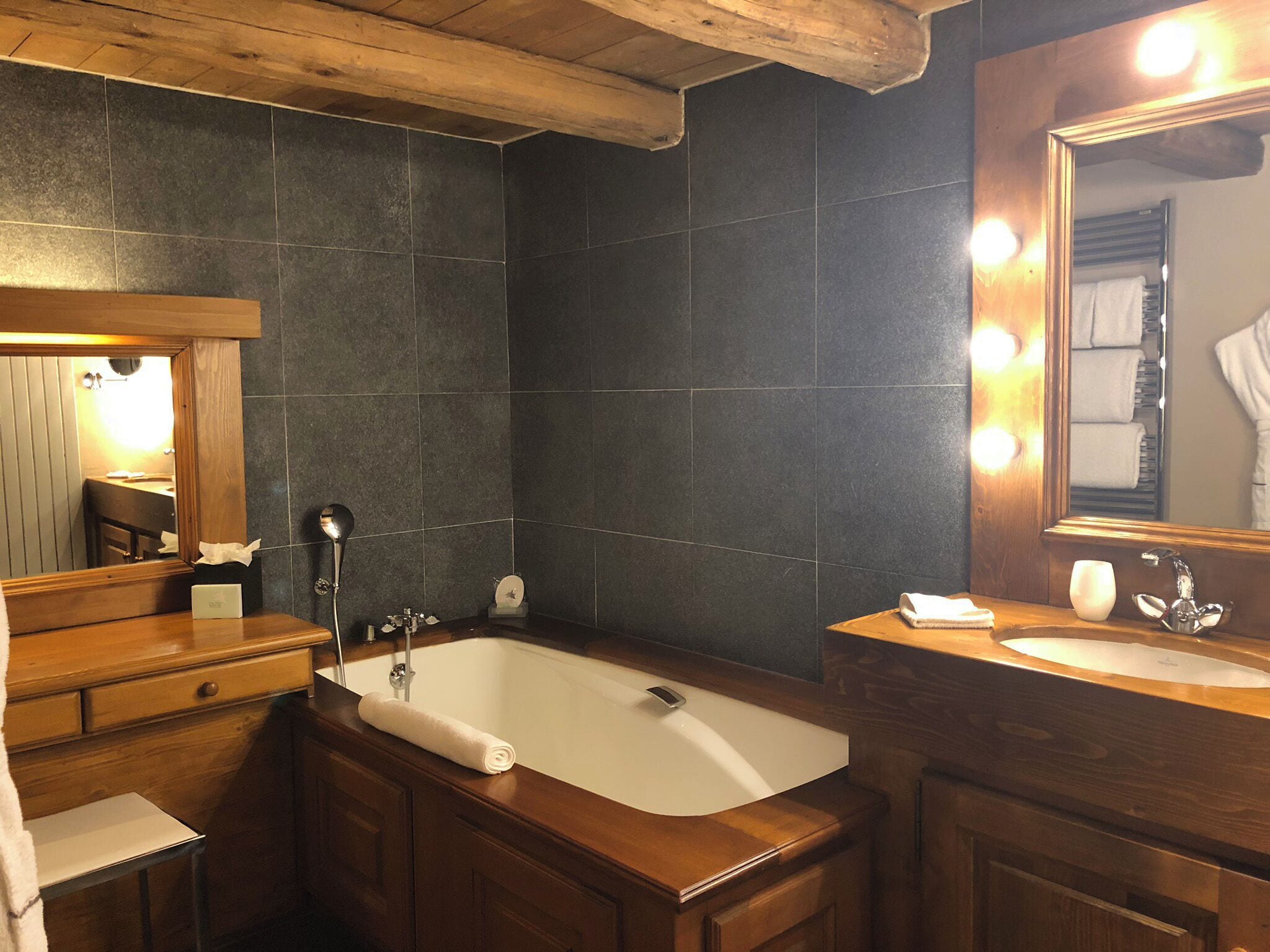 A bathroom with a wooden vanity, a large bathtub, and a mirror with lights.