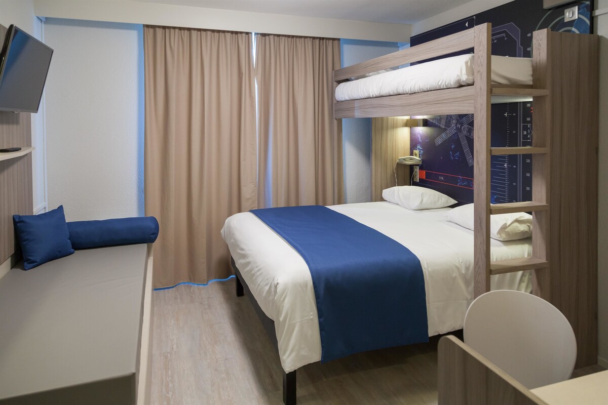 A hotel room with a bunk bed, a desk, a chair, a TV, and a window with curtains.