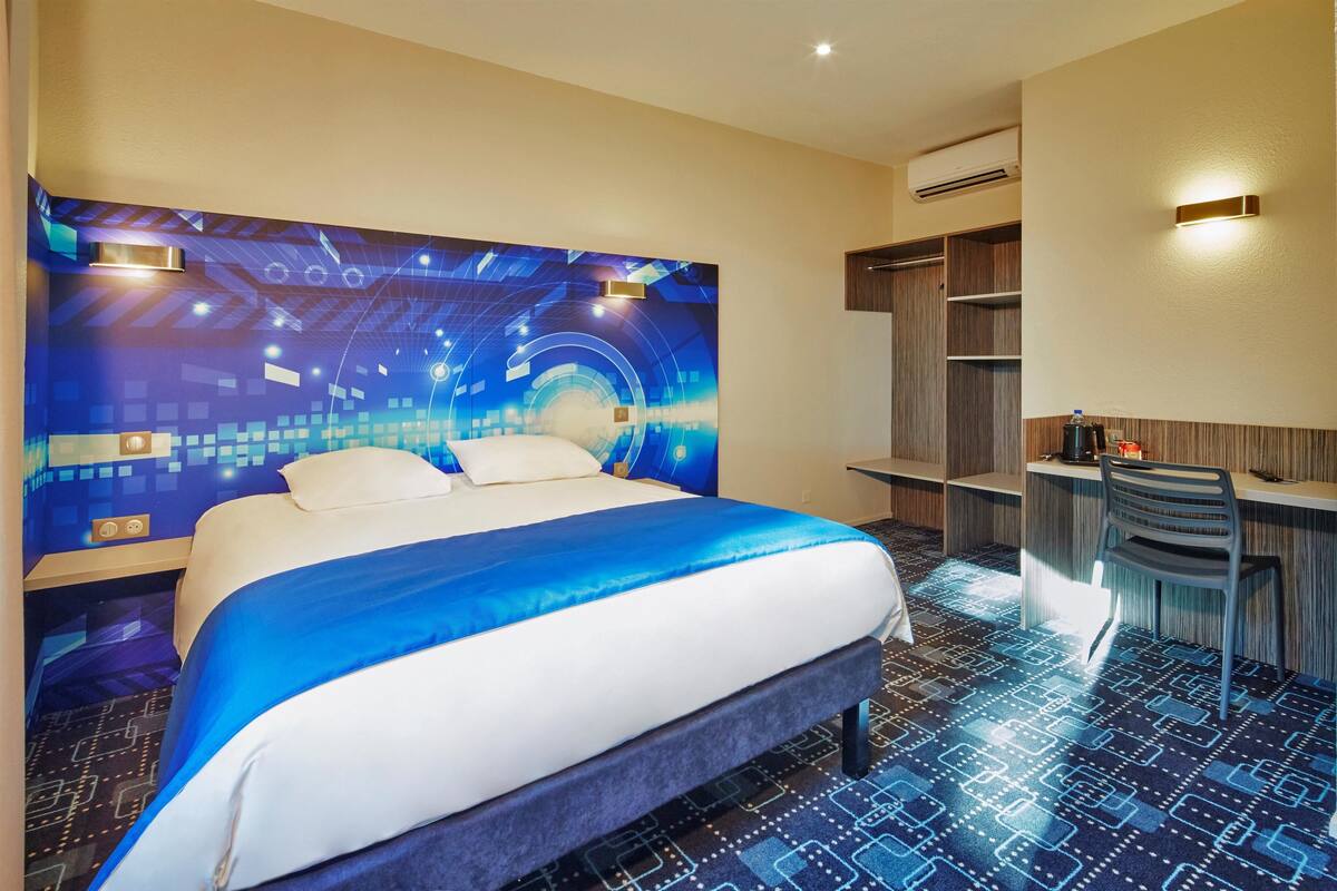 Ein Hotelzimmer mit einem großen Bett, einem Schreibtisch und einem Stuhl. Eine Wand zeigt ein blau-weißes Design, und die Decke ist mit Einbauleuchten versehen.