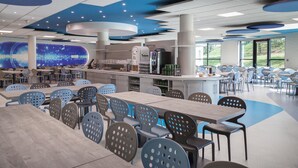 Eine geräumige Cafeteria mit mehreren Tischen und Stühlen, einem zentralen Servicetheke und großen Fenstern mit Blick ins Freie.