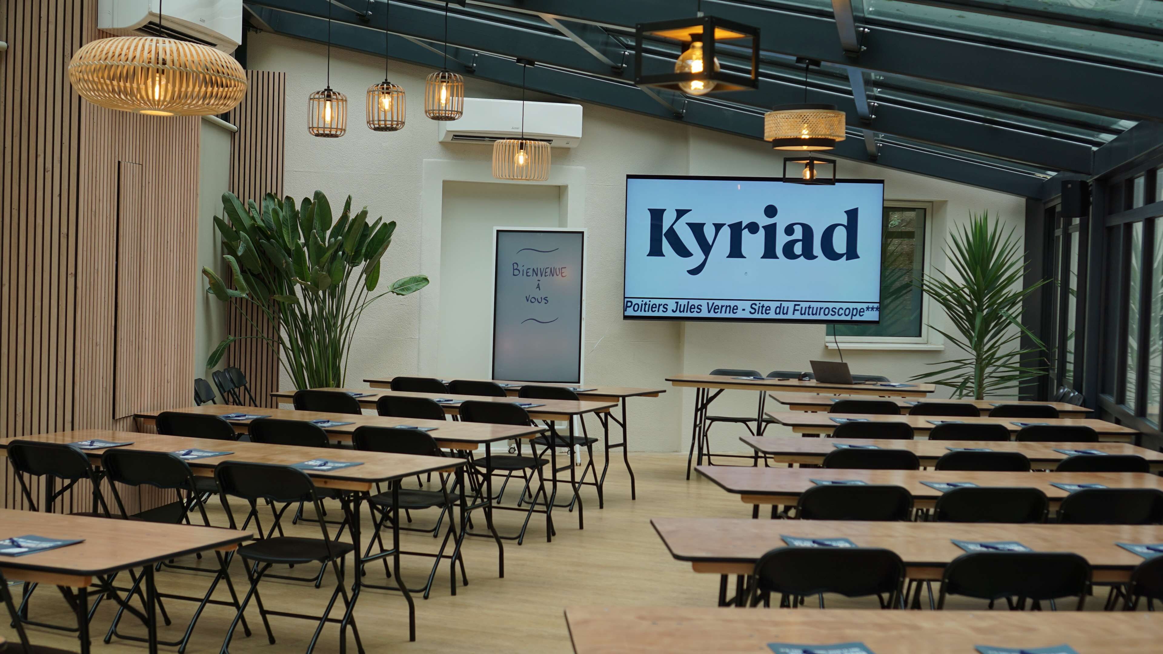 Un salón de conferencias con mesas y sillas, una pantalla grande mostrando 'Kyriad' y un cartel que dice 'Bienvenue à vous'.