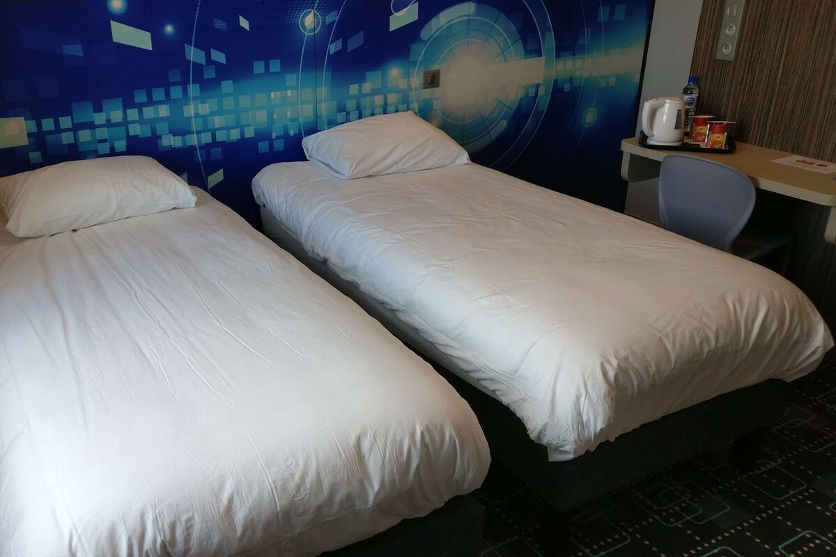 Ein Hotelzimmer mit zwei Einzelbetten, einem Schreibtisch mit Stuhl, einer Kaffeemaschine und einer Wand mit digitalem Design.