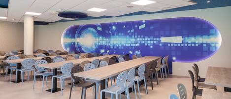 A conference room with multiple tables and chairs, a large digital display on the wall, and a ceiling with recessed lighting.