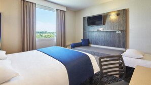 Ein Hotelzimmer mit einem großen Bett, einem Schreibtisch, einem Stuhl, einem an der Wand montierten Fernseher und einem Fenster mit Vorhängen.