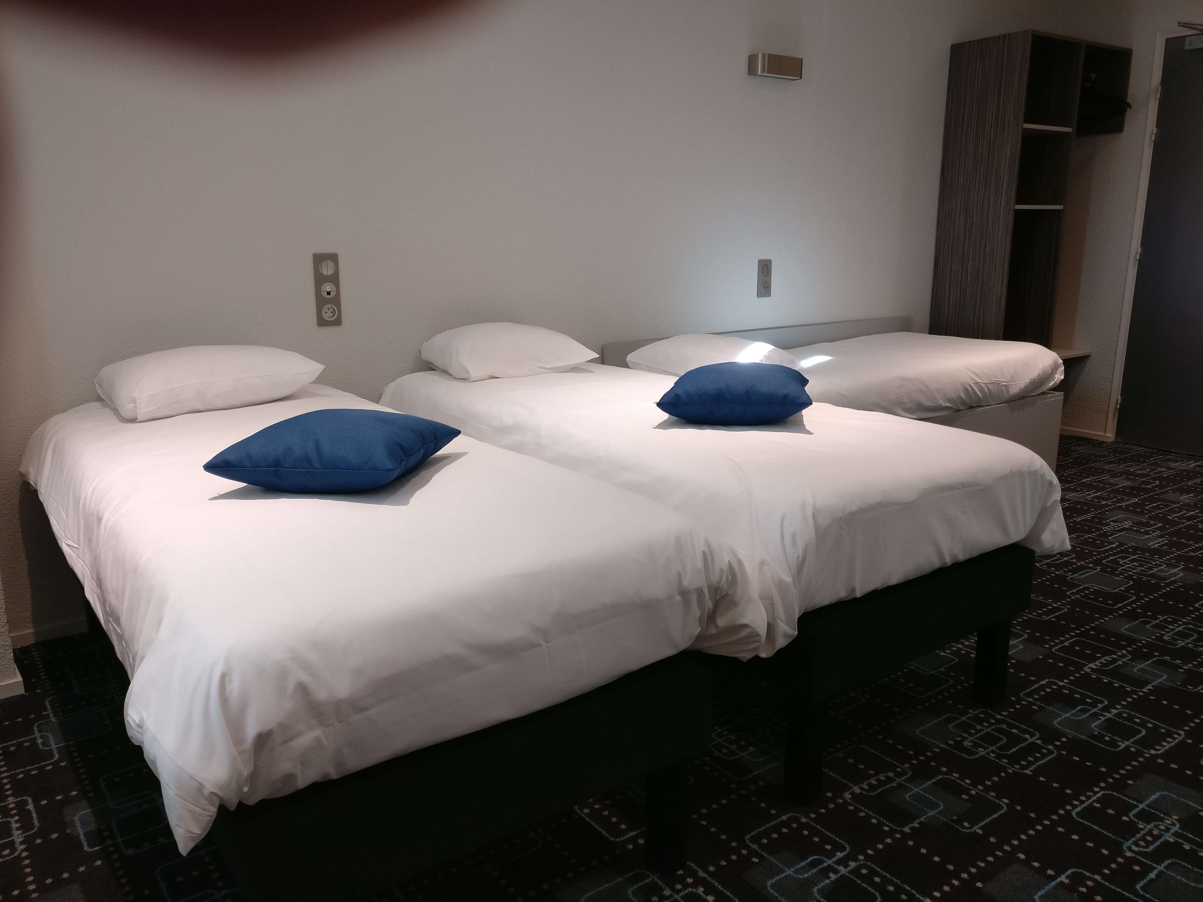 Une chambre d’hôtel avec deux lits, chacun doté de draps blancs et d’un oreiller bleu. Une petite lampe fixée au mur se trouve au-dessus des lits, et une armoire de couleur sombre est placée sur le côté droit.