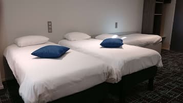 Una habitación de hotel con dos camas, cada una con ropa de cama blanca y un almohadón azul. Hay una lámpara pequeña de pared sobre las camas y un armario oscuro a la derecha.