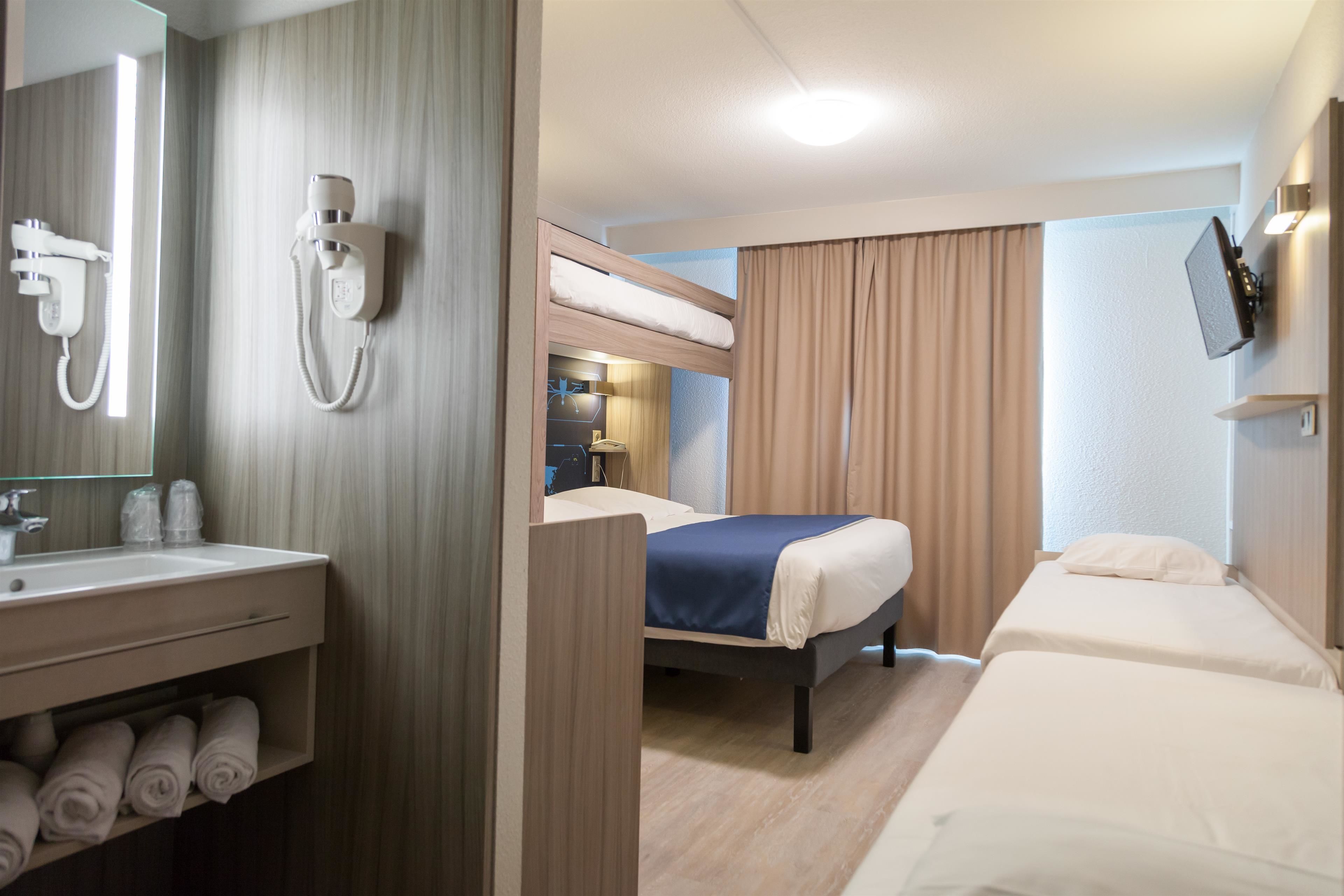 Una camera d'albergo con un letto a castello, un letto singolo, un bagno con lavabo e specchio, e una TV fissata al muro.