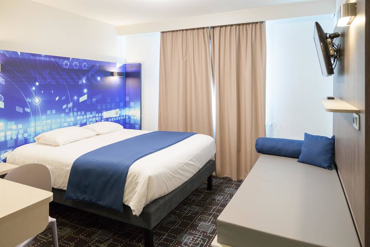 Ein Hotelzimmer mit einem großen Bett, einem kleineren Bett, einem Schreibtisch und einem an der Wand befestigten Fernseher.