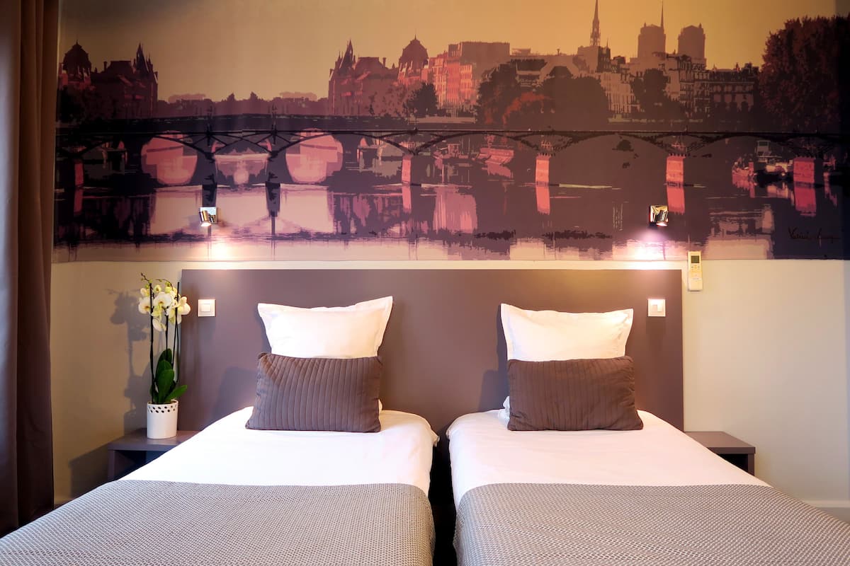 A hotel room with two beds, a cityscape mural on the wall, a potted orchid, and a nightstand with a lamp.