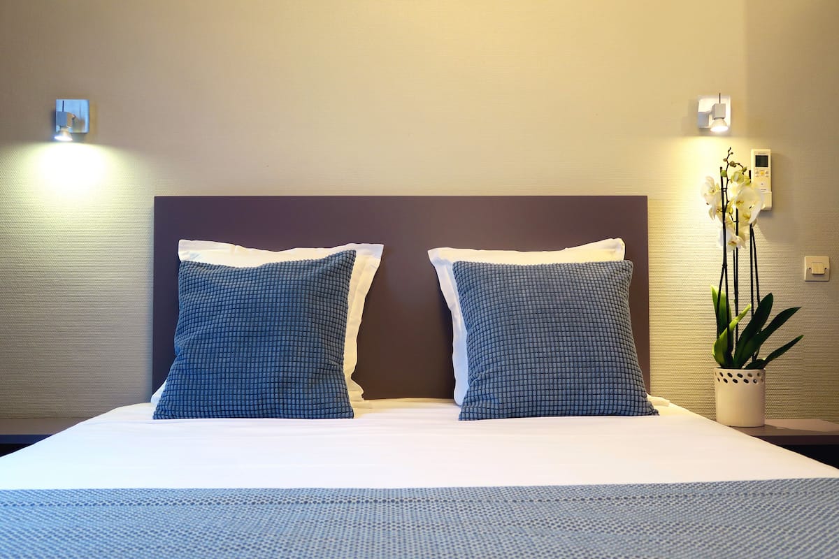 A neatly made bed with two pillows, a bedside plant, and wall-mounted lights.