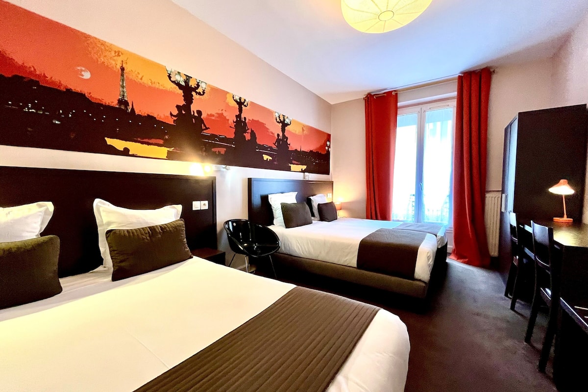 A hotel room with two beds, a desk, and a chair. There is a painting on the wall and a window with red curtains.
